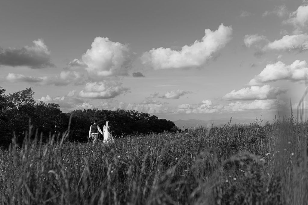 Black and white image of a bride and groom walking through a field of wildflowers.
