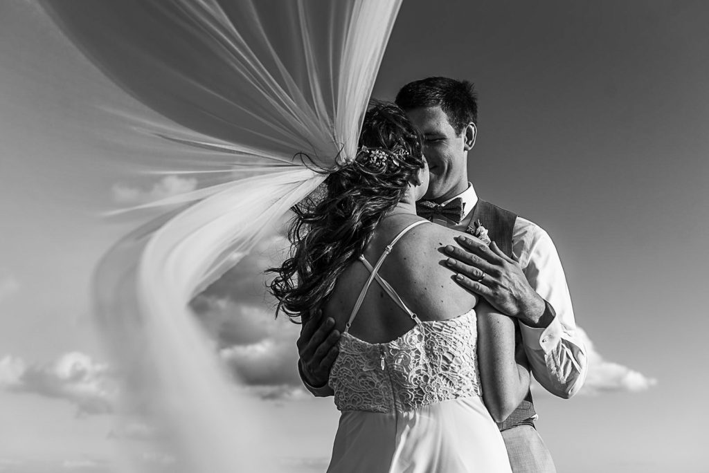 Bride and groom hugging while veil flies in the background in black and white