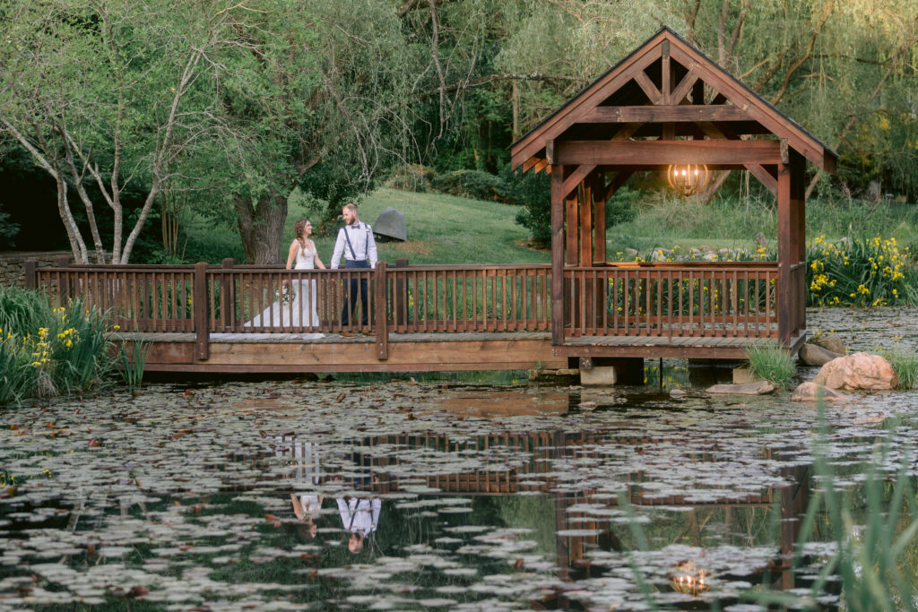 The bride and groom walk together on a bridge over a pond of lilly pads.