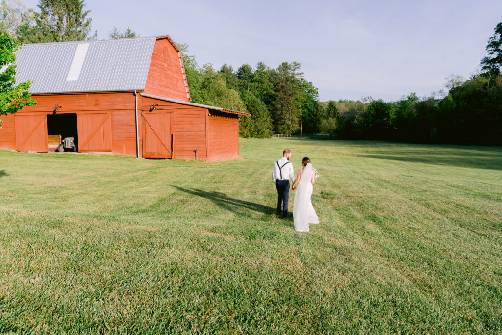 The bride and groom walking together through a field behind a red barn.